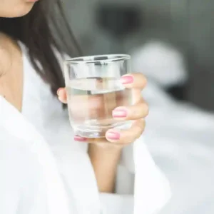 A close-up of a woman in a white robe holding a glass of clear water, showcasing her pink manicure.
