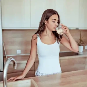 A woman in a white tank top drinks water from a glass while standing at a kitchen counter with wooden decor.