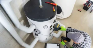 A technician in a gray cap and green gloves works on a cylindrical water heater, using tools and inspecting pipes.