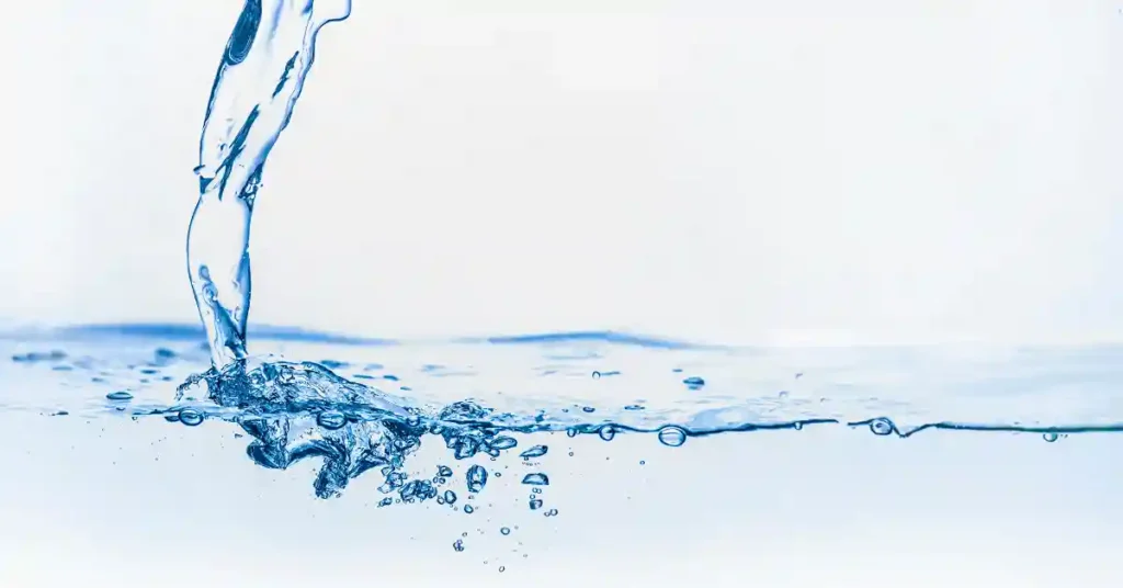 A stream of clear blue water splashes into a serene body of water, creating ripples and tiny bubbles against a white background.