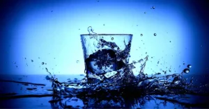 A clear glass splashes water against a blue backdrop, creating dynamic droplets and ripples around it.