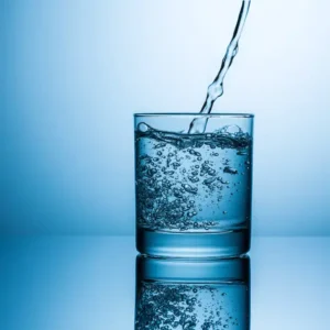 A clear glass fills with sparkling water, creating bubbles, against a soft blue background with a reflective surface.