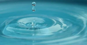 A close-up of water droplets splashing into a serene, blue surface, creating circular ripples in the tranquil water.