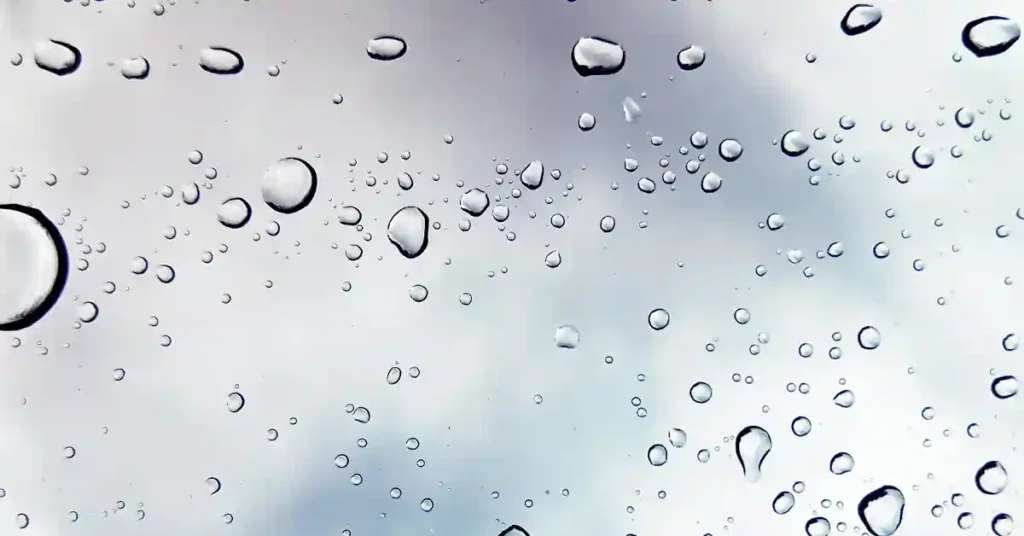 Close-up of raindrops on a transparent surface, with blurred gray clouds in the background creating a soft, soothing atmosphere.