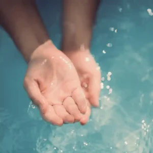 Two gently cupped hands are submerged in clear blue water, with droplets glistening on the surface.