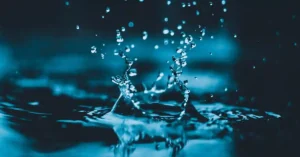 A close-up of a water droplet creating a splash in a deep blue pool, with droplets spraying around, capturing a moment of fluid motion.