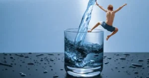 A boy in swim trunks leaps into a large glass filled with splashing water, surrounded by droplets on a dark surface.