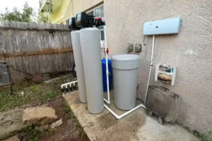 A water filtration system featuring two large tanks, a blue barrel, and connected pipes, set against a weathered fence.