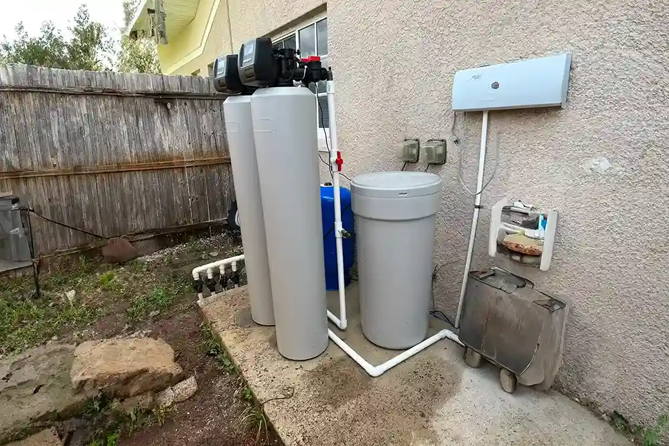 A water filtration system featuring two large tanks, a blue barrel, and connected pipes, set against a weathered fence.